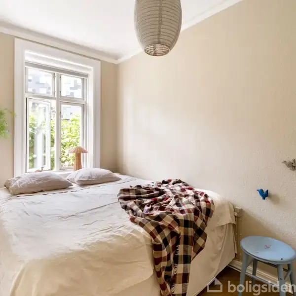 En dobbeltseng står centreret i et lyst soveværelse med beige vægge, opredt med puder og en ternet plaid. Et vindue bringer naturligt lys ind, ved siden hænger en papirlampe.