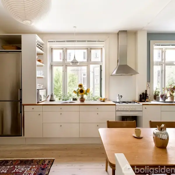 Køkken med køleskab, komfur og hvid bordplade. Et bord med en vase og krus står i midten. Store vinduer slipper lys ind, omgivet af planter.