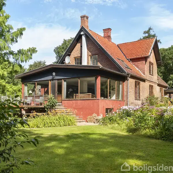 Murstenshus med rød tagsten og stor veranda omgivet af frodig have med græsplæne og træer under en klar blå himmel.