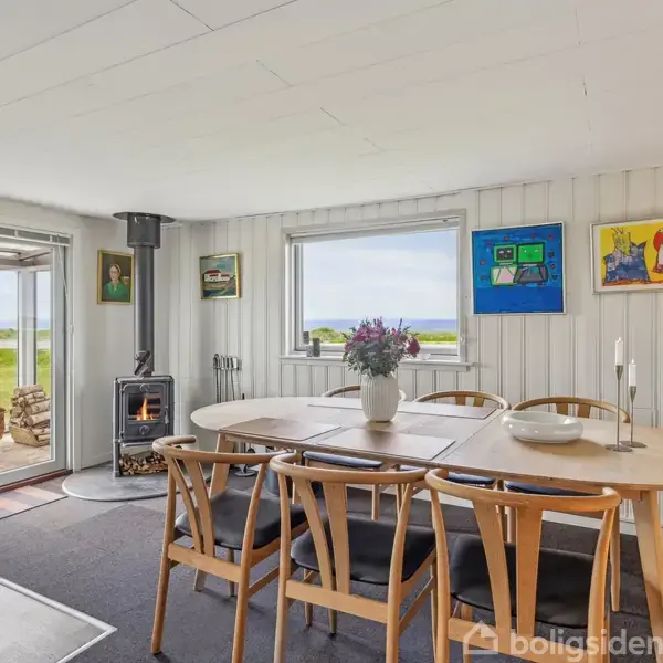 Et spisebord med stole står i et lyst rum med hvidmalede vægge. En vase med blomster pynter bordet, mens en brændeovn brænder i hjørnet. Billeder hænger på væggen. Udenfor ses en terrasse og grønne marker.