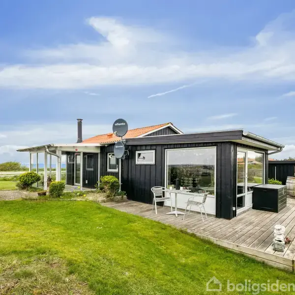 Et sortmalet sommerhus med store vinduer står på en træterrasse. På terrassen er der en siddegruppe. Huset er omgivet af en grøn græsplæne under en klar blå himmel.