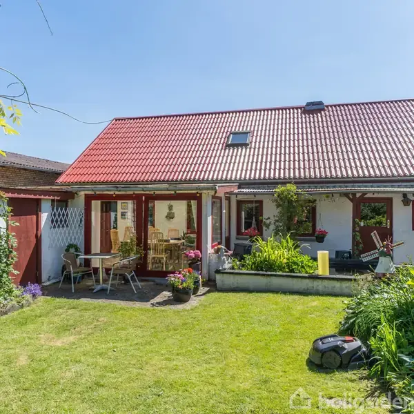 Et lille hus med rødt tag står i en solrig have. En terrasse med havemøbler er foran huset, omgivet af grønne planter og blomster, mens en plæneklipper er på græsplænen.