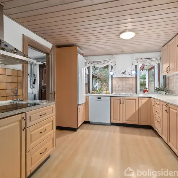 Køkken med træfarvede skabe og ståludstyr. Køleskab, ovn og opvaskemaskine ses langs siderne. Brede vinduer i hjørnet giver udsigt til haven. Lysebrun flisevæg over køkkenbordet.