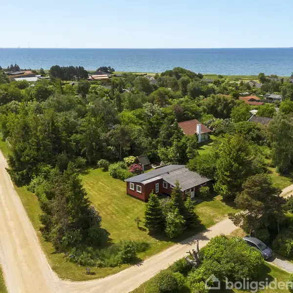 Et rødt hus med gråt tag omgivet af frodig vegetation. Huset står på en stor grøn grund med grusveje omkring sig. I baggrunden ses havet under en blå himmel.