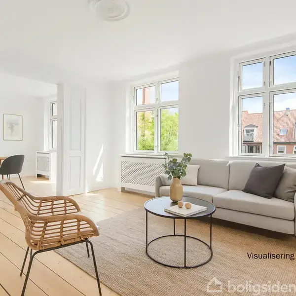 En stue med en grå sofa står ved et rundt bord med planter, placeret på et lyst trægulv. Store vinduer giver naturligt lys, og en lænestol står ved siden af. Tekst: "Visualisering".