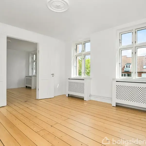 Lyst rum med trægulve og hvide vægge, to vinduer med udsigt til røde tegltage, og opvarmningsradiatorer under vinduerne. En åben dør fører til et tilstødende rum.