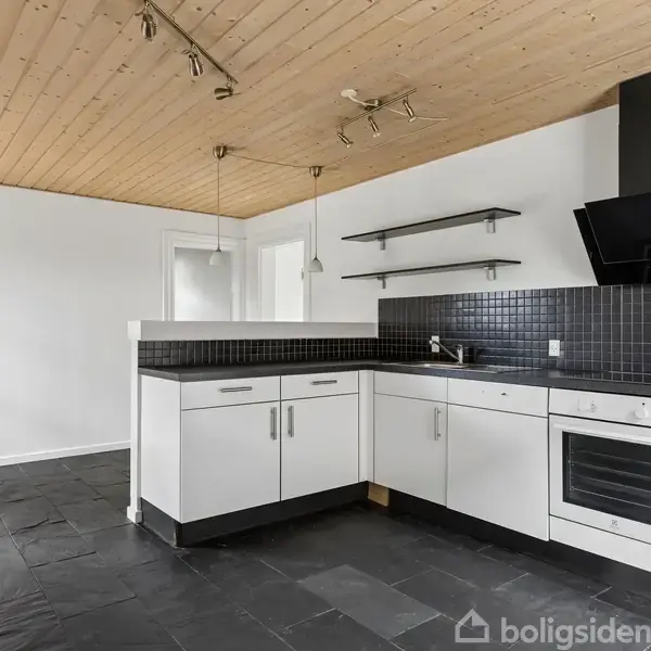 Moderne køkken med sorte fliser som gulv og backsplash, hvide skabe, indbygget ovn og emhætte. Træloft med spots. Dør fører til næste rum med træparketgulv.