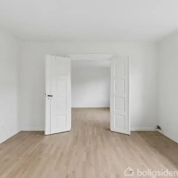 Dobbelt hvide døre står åbne i et rum med lyst trægulv. Sollys trænger ind fra et vindue til højre, mens en radiator er placeret under vinduet.