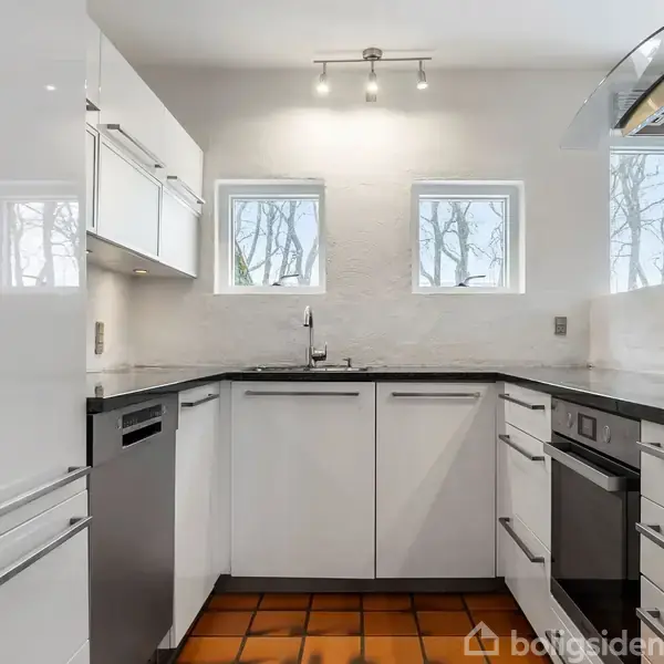 Et moderne køkken med hvide skabe og sort bordplade. Komfur og opvaskemaskine til venstre. To vinduer giver naturligt lys. Flisegulv og loftsbelysning fuldender rummet.