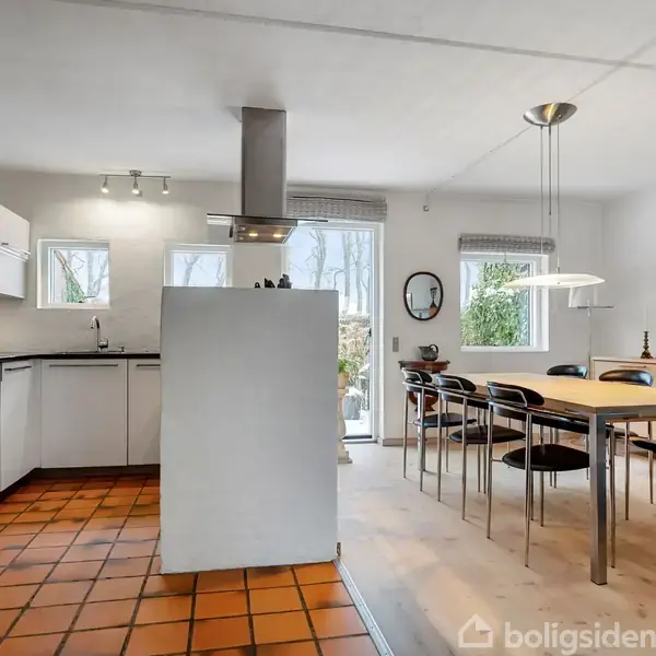 Et moderne køkken forbundet til en spiseplads med et langt træbord og otte stole. Flisegulv skifter til trægulv. Store vinduer giver udsigt til haven.