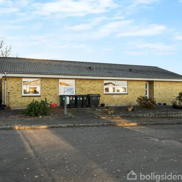 Gulstensbygning med fladt tag står stille ved fortovskant. Foran bygningen ses skraldespande og en trailer. Træer uden blade omgiver området under en blå himmel.