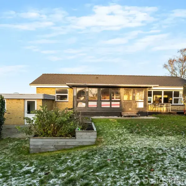 En gul murstensvilla med stor veranda står i en have med sparsom sne og bevoksning. Til højre er et dække og en trailer. Himlen er blå med få skyer.