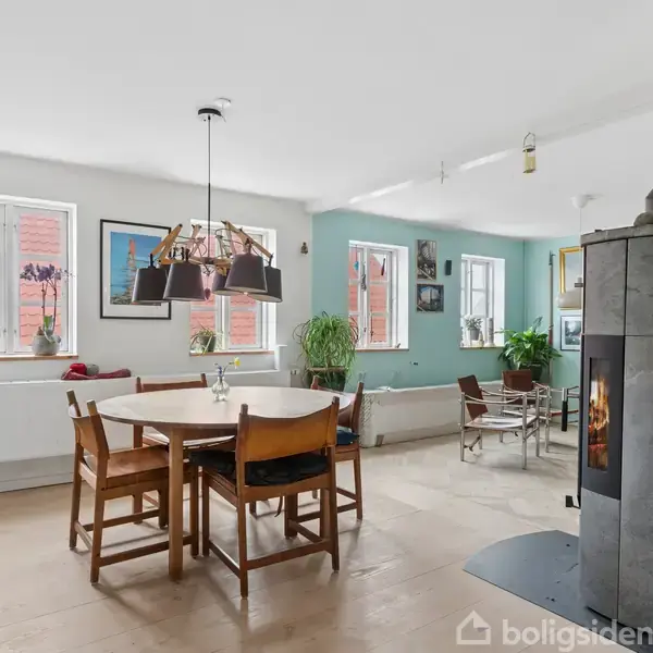 Spisebord med stole står i lyse stue med brændeovn. Store vinduer, planter og kunst på væggene skaber en hyggelig atmosfære. Trappe ses til venstre.