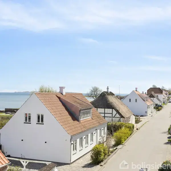 En række hvide huse med røde tage står langs en fredelig vej. Bag husene ses havet i horisonten under en klar, blå himmel.