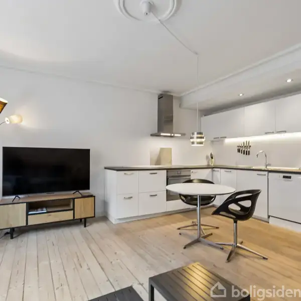 En moderne stue med gråt sofaarrangement står ved et stort vindue. Rummet har trægulv, et rundt spisebord med sort-hvide stole, og der hænger en lampe over bordet. I hjørnet ses et TV og en ur.