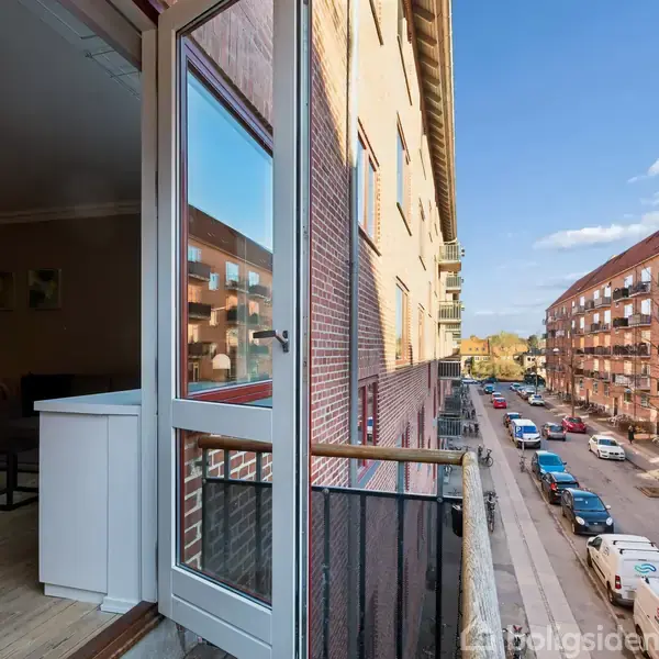 En lys gang med fliser på gulvet og hvidmalet dør. Til højre er en åben døråbning til en stue med pastelvægge og dekoreret med to malerier.