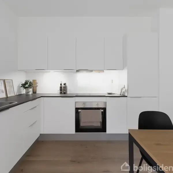 Et moderne køkken med hvide skabe og indbygget ovn under en sort bordplade. Rundt om er der dekorationer og madlavningsredskaber. Til højre står et træbord med stole.