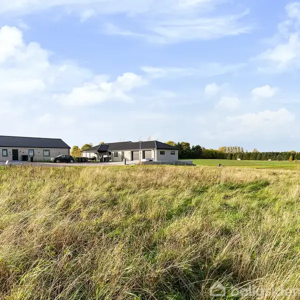 Et moderne hus står stille ved en åben eng med højt græs. I baggrunden ses flere huse, omgivet af grønt landskab og en klar blå himmel med få skyer.