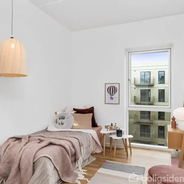 Seng dækket med tæppe og puder står ved væggen i et lyst værelse. Bord med lampe ved siden af vindue med udsigt til lejligheder. Intet synlig tekst.