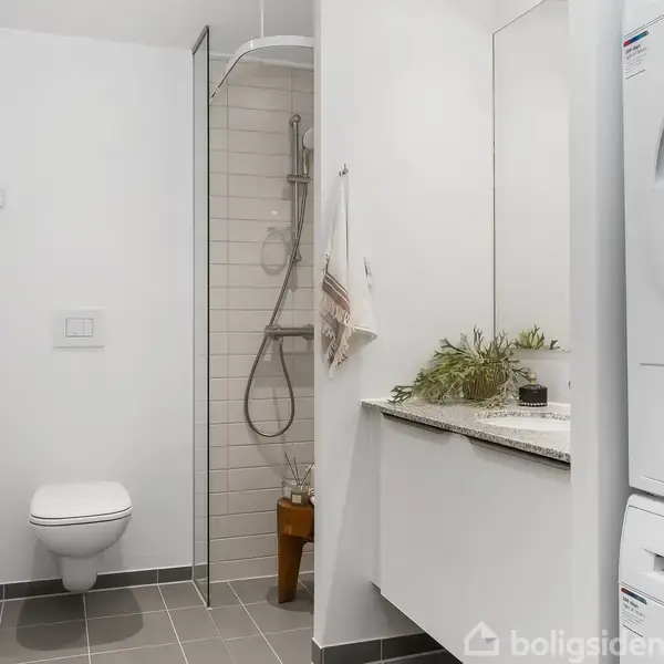 Et moderne badeværelse med væghængt toilet, brusekabine med glasvæg, hvid vaskemaskine og tørretumbler fra Bosch. En vase med planter står på en hvid bordplade ved spejlet.