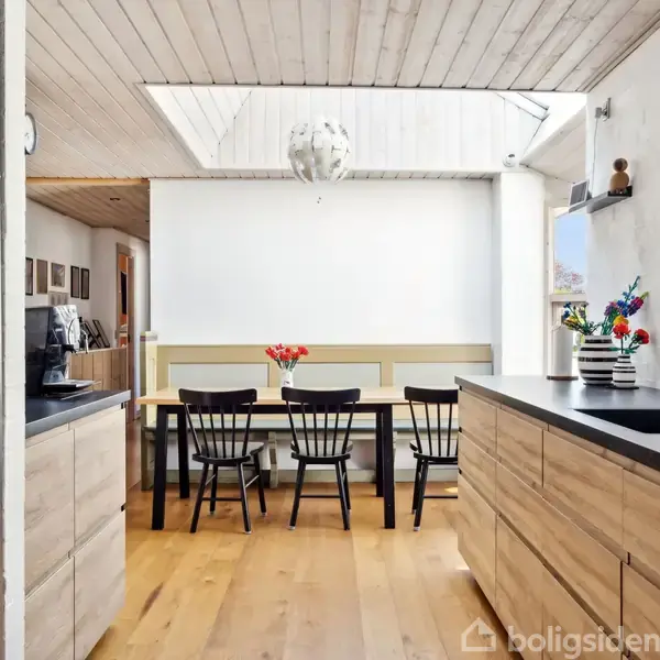 Et moderne køkken med træskabe og sort bordplade. Bord med fire stole og blomsterdekorationer i baggrunden. Loftet har ovenlys, og lyset strømmer ind gennem et vindue.