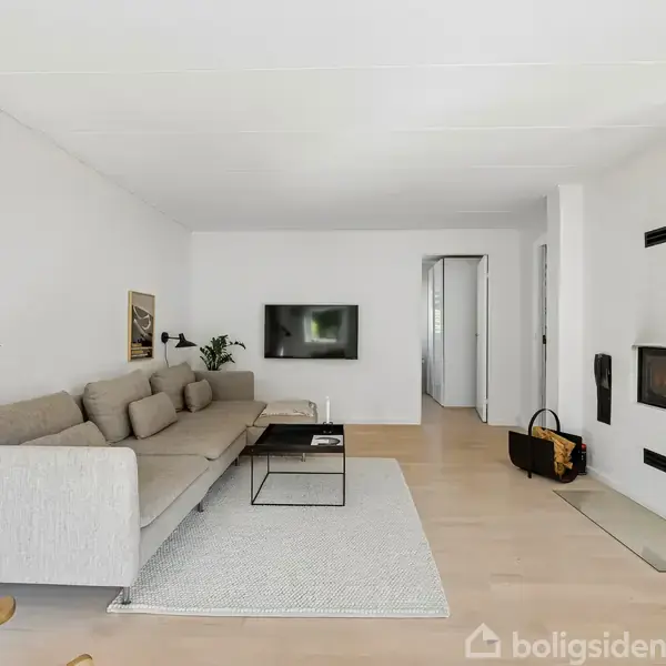 En grå sofa står langs væggen i en minimalistisk stue. Et tv hænger på væggen, og en moderne pejs er indbygget i højre væg. Et lille tæppe ligger foran sofaen.