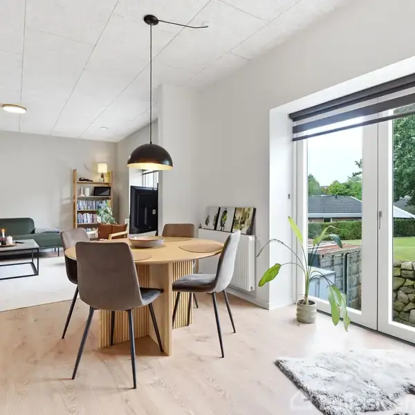 En moderne spisestue med et rundt træbord og stole, oplyst af en hængelampe. Rummet åbner op til en grøn have gennem store glasdøre, med en stue i baggrunden.