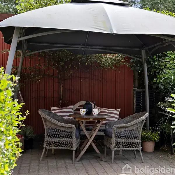 Pavillon beskytter et bord med to kopper og flettede stole. Omkring er der frodigt grønne planter og et rødt hegn, hvilket skaber en hyggelig haveatmosfære.