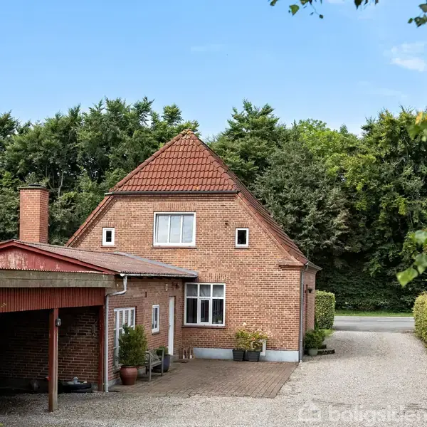 Et rødt murstenshus står stille, omgivet af grusbelagt indkørsel og tæt bevoksning af træer i baggrunden. Carport til venstre, med flisebelagt sti foran hoveddøren.