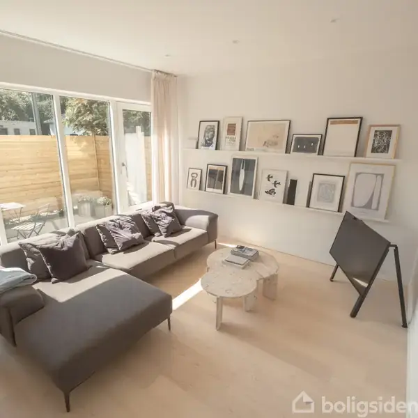 Grå sofa med puder står i en soloplyst stue ved siden af et sofabord. Væghylde med indrammede billeder hænger over et staffeli. Store vinduer giver udsigt til en terrasse.