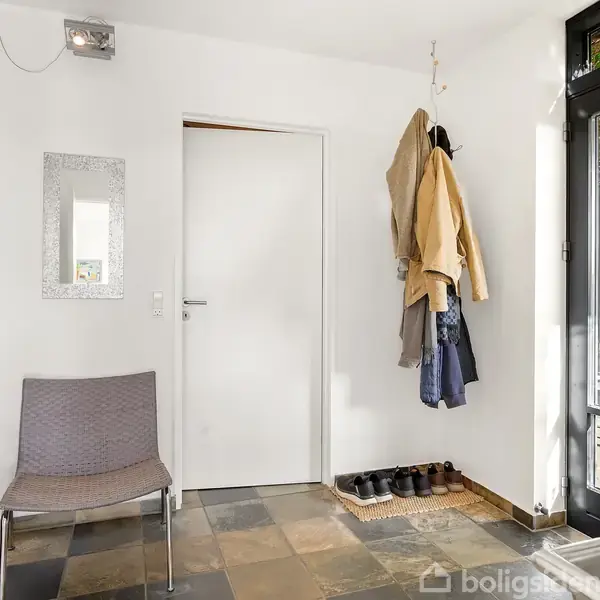 En stol står i en entré med flisegulv. Jakker hænger på en knagerække ved siden af en sko- og måttestation. En glasdør fører ud til en terrasse.