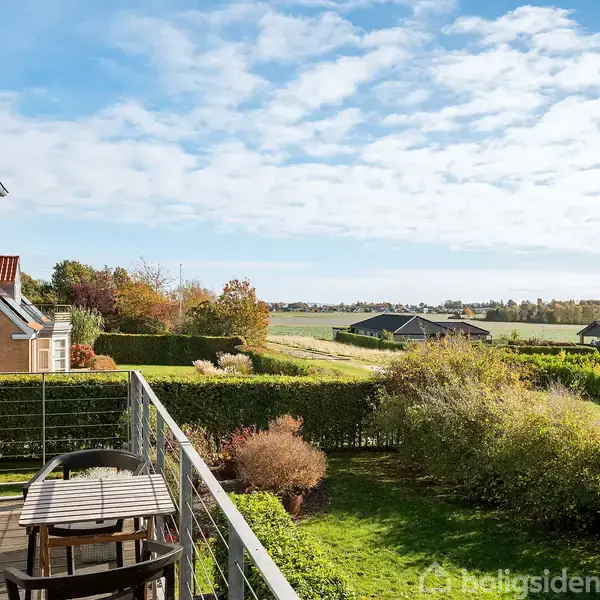 En træterrasse med et bord og to stole står i forgrunden. Terrassen har udsigt over grønne marker og et parcelhuskvarter under en blå himmel med lette skyer.