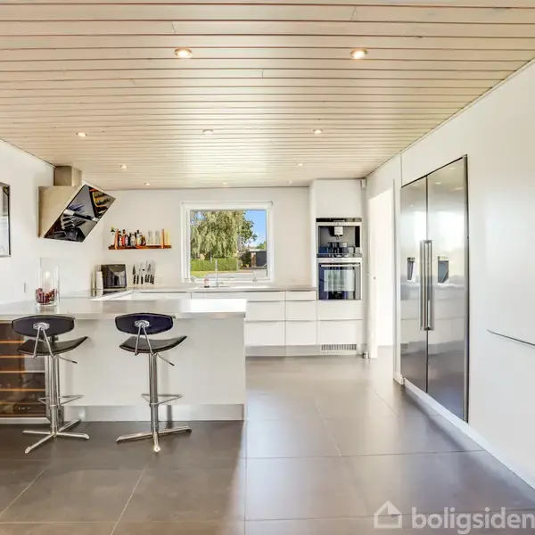 Køkken med to moderne barstole ved en bardisk, køkkenapparater indbygget i hvide skabe, og et vindue med udsigt til en grøn have. Klinkegulv og træloft.