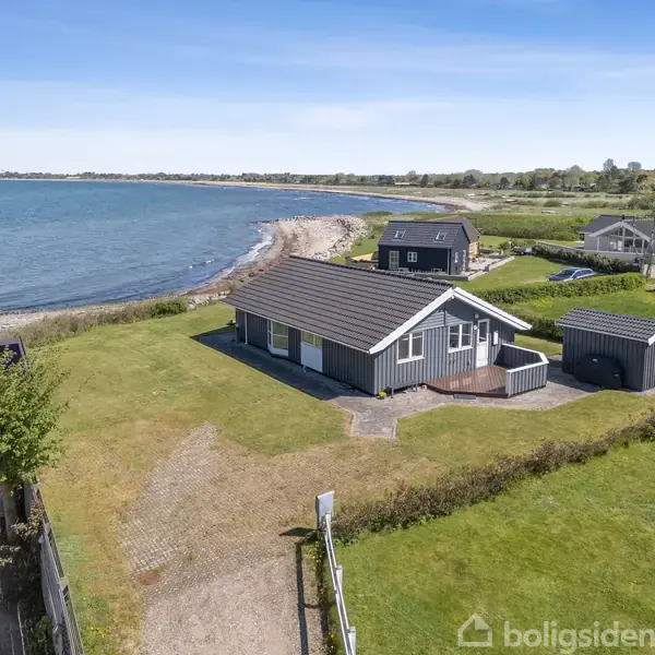 Et gråt sommerhus med et sort tag står tæt ved en kystlinje ved havet. Det ligger omgivet af en grøn græsplæne, og der er også et lille skur ved siden af.