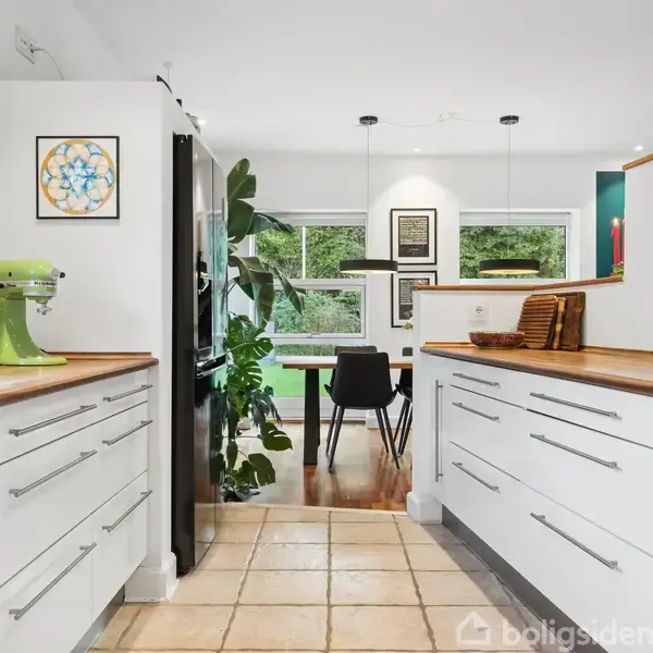 Køkken med hvide skabe og træbordplader, en grøn mixer står på bordpladen. Spiseplads i baggrunden med sort bord og stole, omgivet af store vinduer og grønne planter.