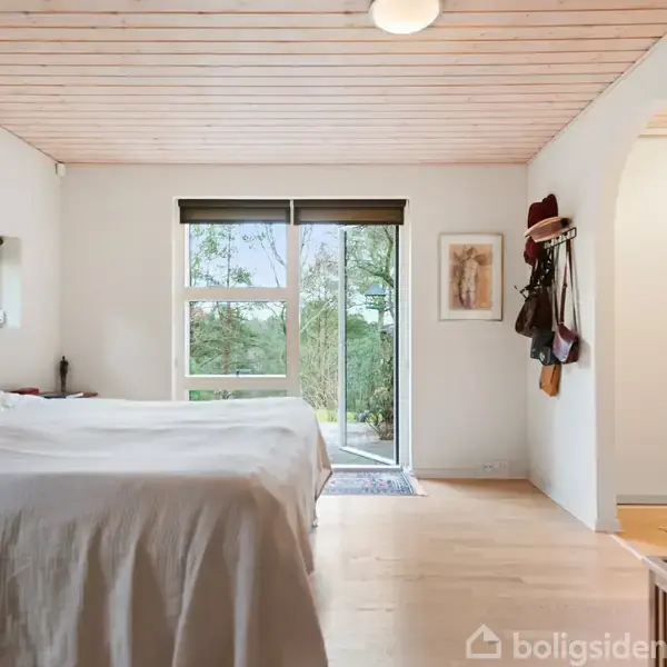 Seng dækket med hvidt sengetøj står i et lyst værelse med træloft. Store vinduer viser en grøn udsigt. En gang til højre har tøj og tasker hængende.