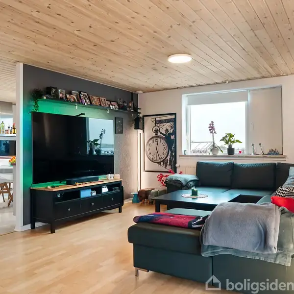 Sort sofa arrangeret i hjørnet foran et stort tv i en stue med lyst trægulv og træloft. Åben forbindelse til køkkenet med spisebord. Vindue med planter bagved.