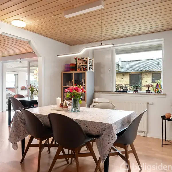 Et spisebord med blomster i vase står dækket med dug i et lyst køkken/alrum. Omgivet af stole, træloft og udsigt til en baggård gennem vinduet.