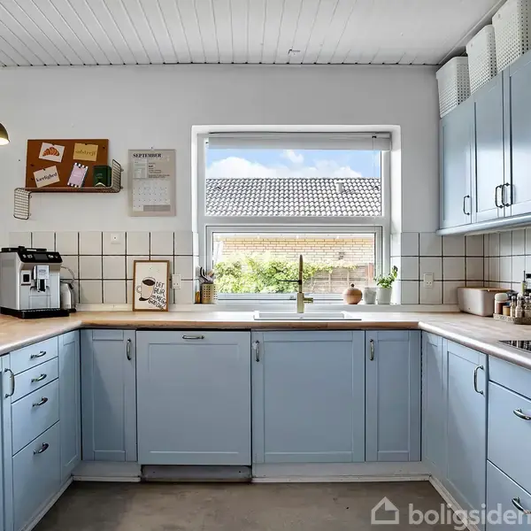 Køkkenet har blå skabe og en træbordplade. Kaffemaskine, knive og planter står på bordet. Vindue med udsigt til tag og himmel. Korktavle hænger over kaffemaskinen.