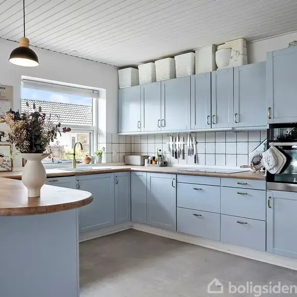 Blå køkkenskabe med hvide fliser over bordpladen, ovn indbygget i skabet. Køkkenø med vase og lamper hængende over. Vindue tillader naturligt lys ind. Ingen tekst synlig.