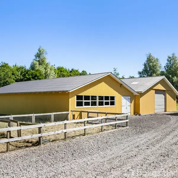 Gul staldbygning med stor port og flere vinduer, beliggende ved en grusvej omgivet af grøn skov og hegn.