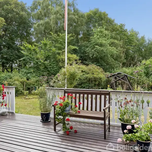 Bænk står på træterrasse omgivet af blomster i krukker. Baggrunden består af frodig have med grønne træer og et hvidt stakit. Flisebelagt sti fører til en flagstang.