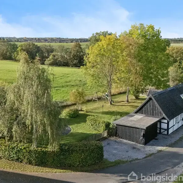 Sort-og-hvid bygning står stille ved landevej, omgivet af grønne træer og marker. Sollys belyser scenen, og der er en gårdsplads ved siden af huset.