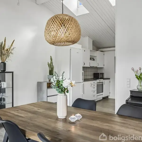 Spisebord i træ med vase, blomster og stearinlys. Hængende fletlampe over bordet. I baggrunden er et moderne køkken og klaver. Skabe, glas og dekorationer ses omkring rummet.