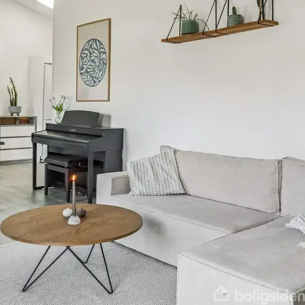 Lysegrå sofa med puder står i en moderne stue. Rundt sofabord har tændte stearinlys. Langvæggen har hylde med planter, mens et klaver står i baggrunden ved spisebordet.