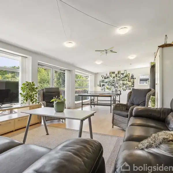 Stue med sorte lædersofaer omkring et sofabord, udsigt til have gennem store vinduer. TV står på skænk, væg dekoreret med billeder. Grøn væg i baggrunden.