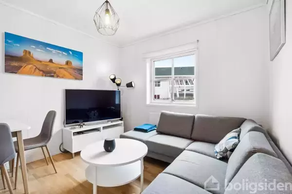 Gråt hjørnesofa-arrangement står foran et stort fladskærms-tv. Værelset har et minimalistisk design med hvidmalede vægge og et farverigt kunstværk. En lille rund sofa bord står i midten.