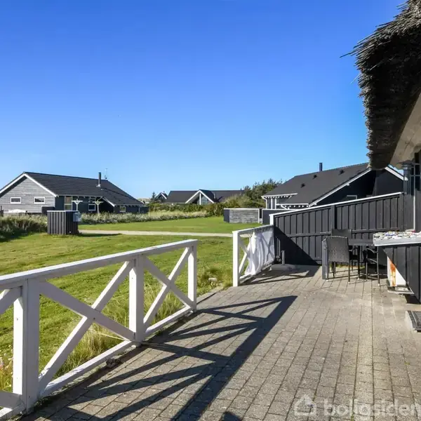 Terrasse foran sort træhus med stråtag, stille ved det grønne græsareal med flere huse i baggrunden, under en klar blå himmel.