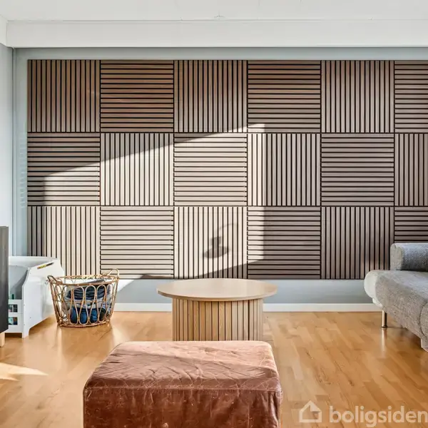 Sofa står i en moderne stue med trægulv og en dekorativ trævæg. Ved siden af er et lavt rundt bord og en brun puf. En flettet kurv står i hjørnet.