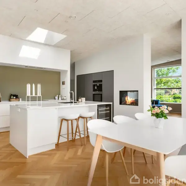 Køkken med hvidt spisebord og barstole; en moderne indretning med åbent design. I baggrunden ses en indbygget pejs og store vinduer, der giver udsigt til grøn have.